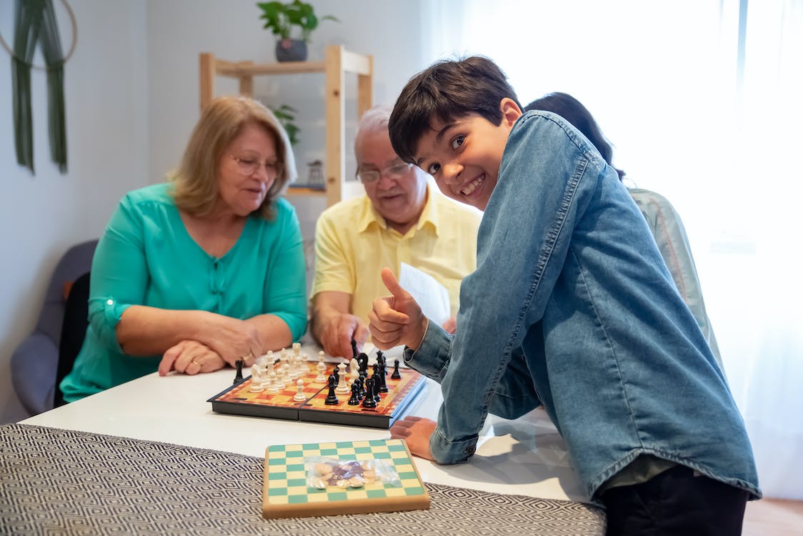 A child showing a thumb up while grandparents are playing chess behind him